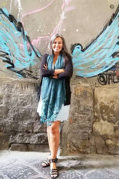 Lupita Eyde-Tucker smiles while crossing her arms in front of a wall with a winged graffiti
